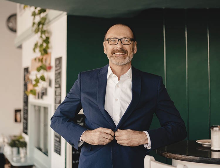 Confident mature businessman in a cafe buttoning his jacket Peltier Financial